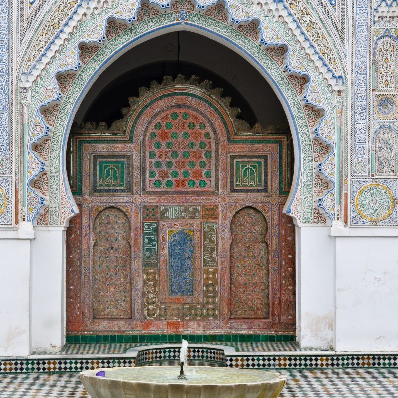 Beautiful mosaic tiled archway over huge wooden doors with a bowl fountain in front.